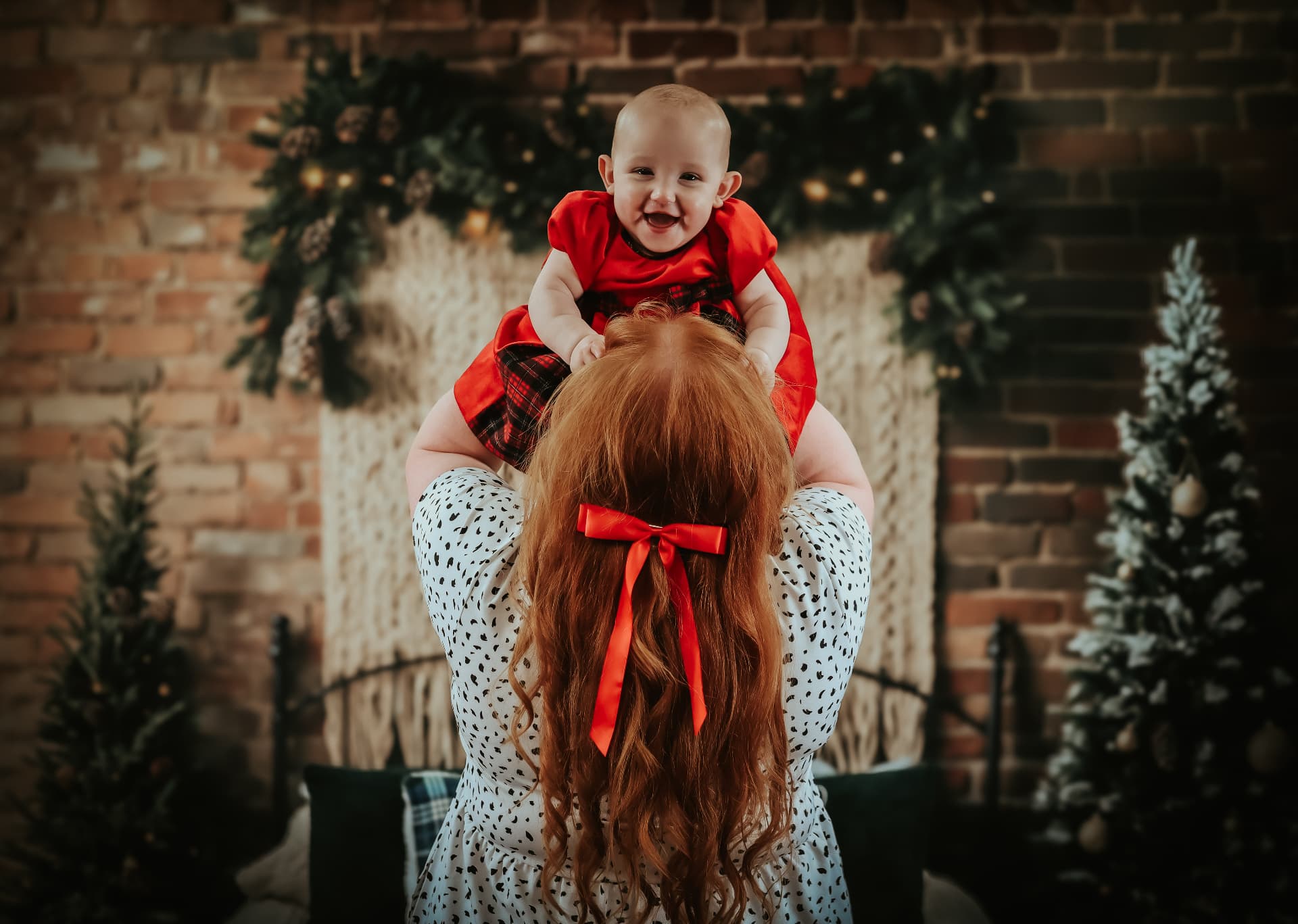 Mother lifting a laughing baby in a red tartan dress against a Christmas garland backdrop
