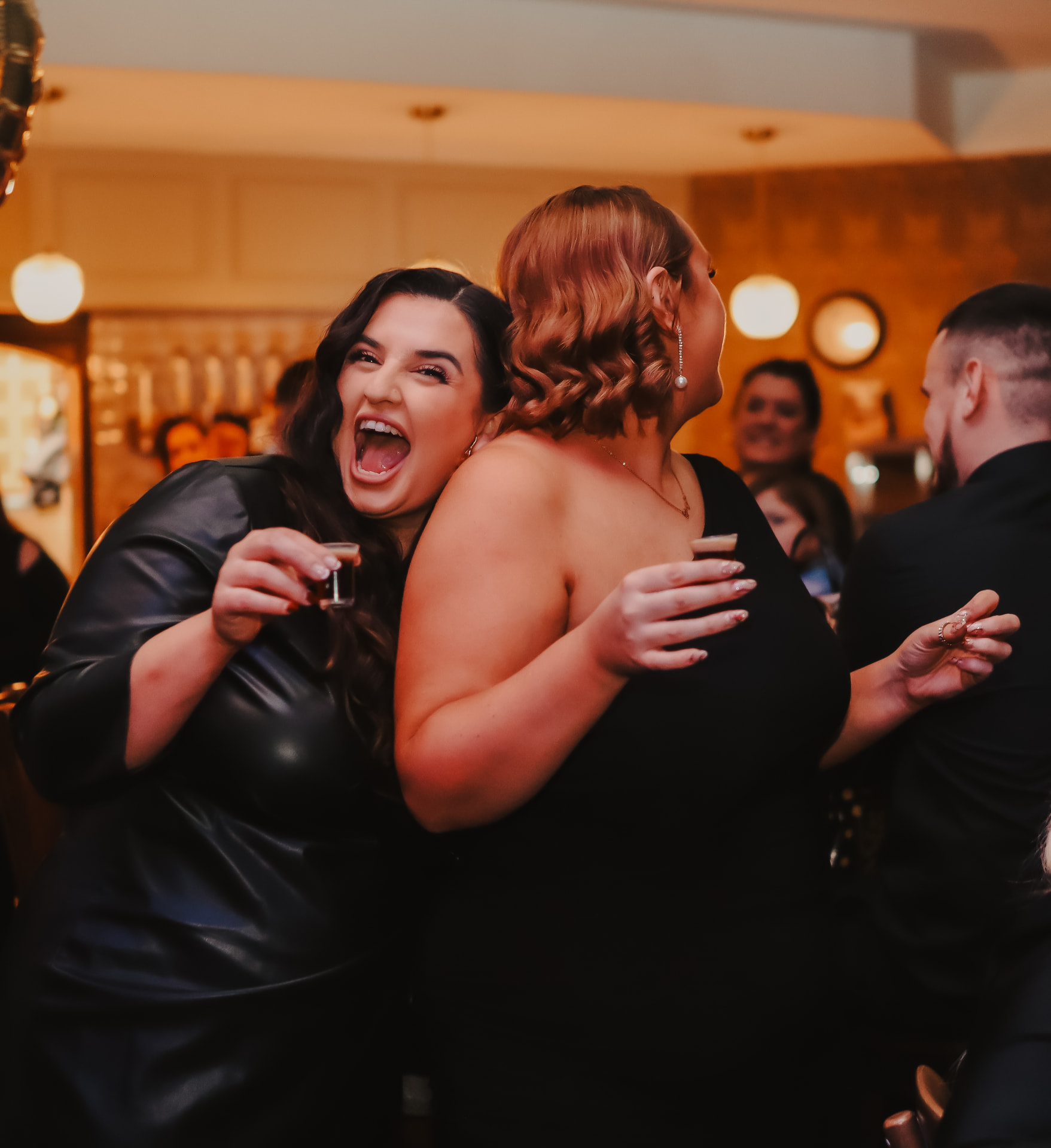 Two women laughing together over drinks at a lively evening celebration