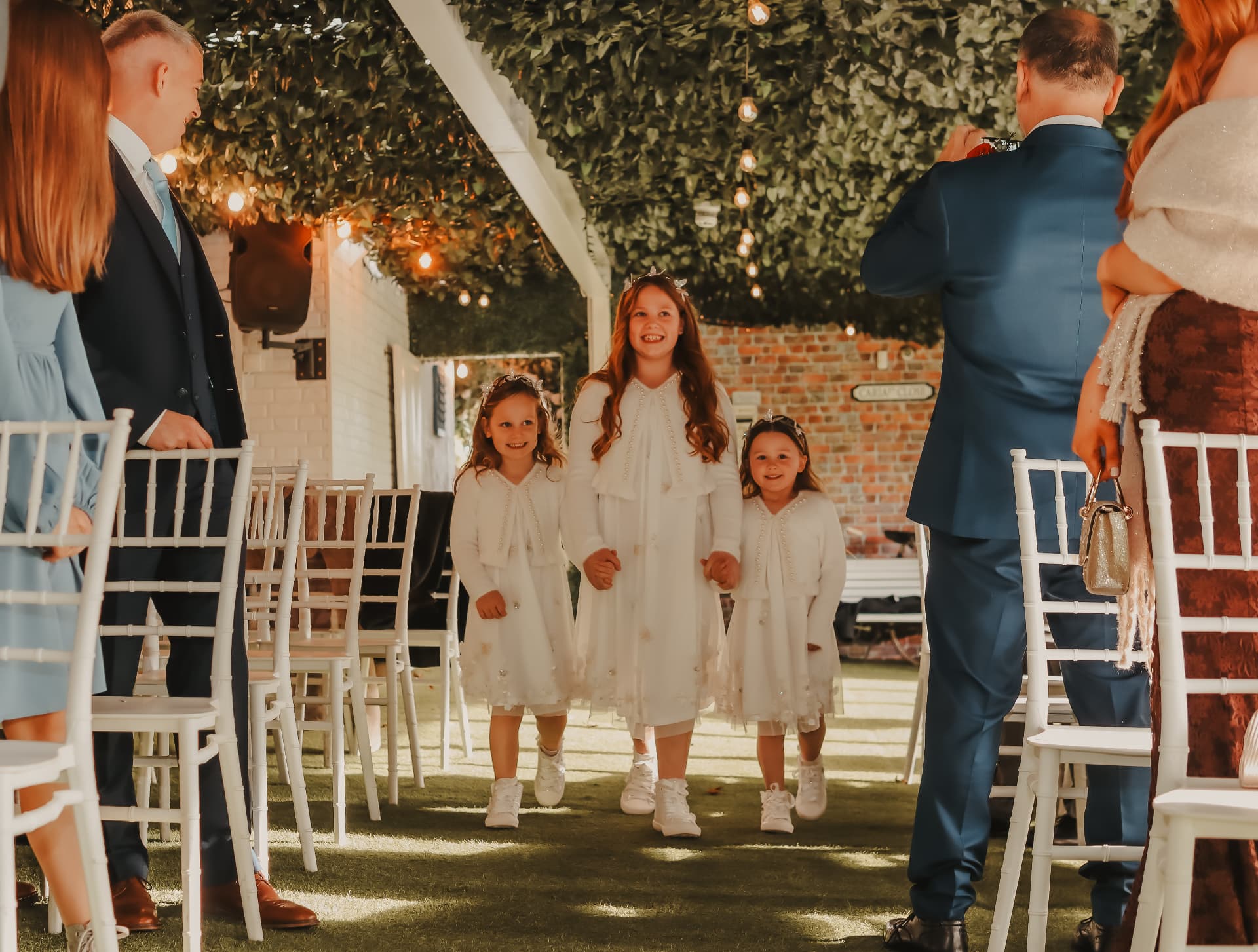 Three flower girls walking down the aisle beneath a foliage canopy