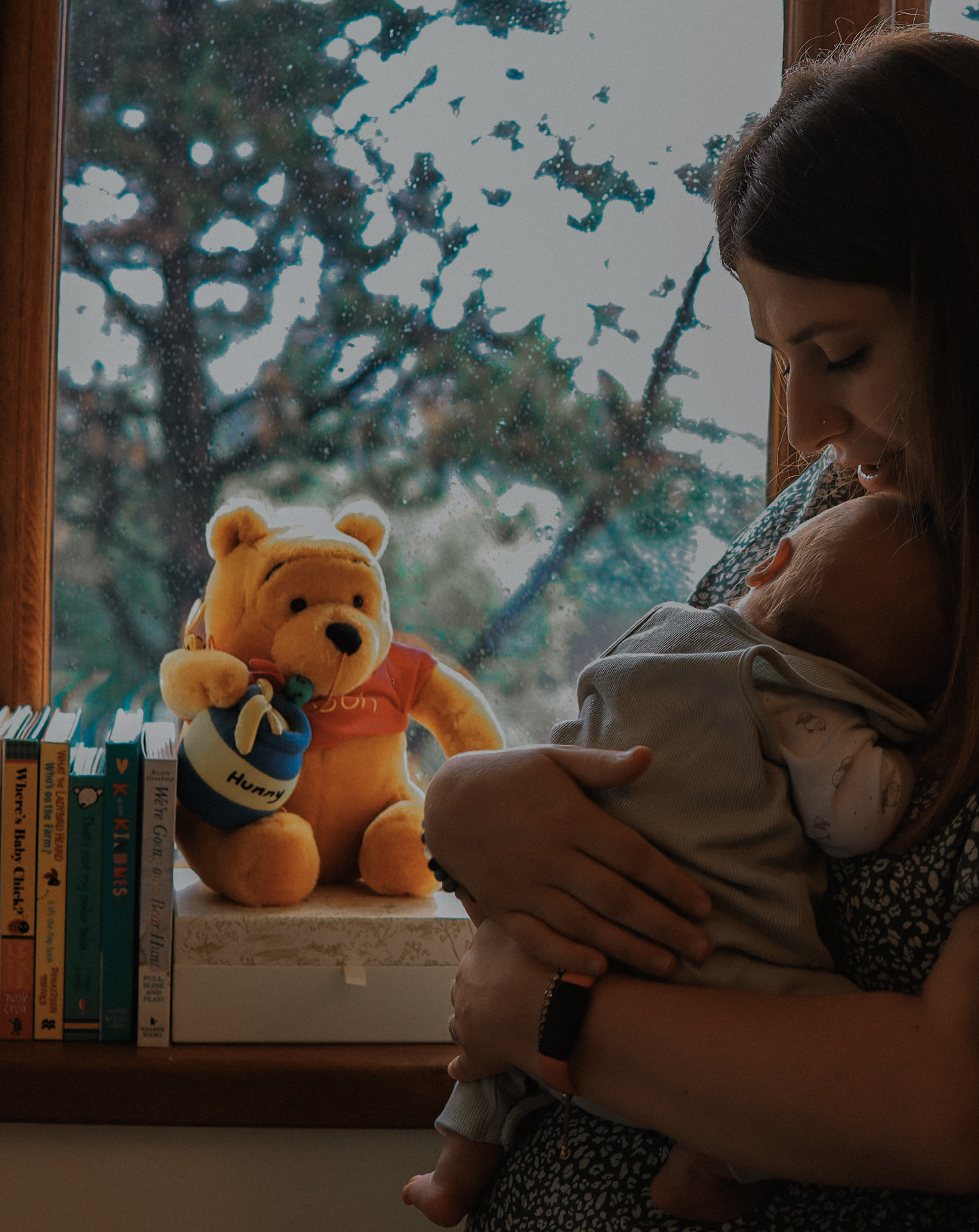 Mother holding her newborn beside a sunlit window with a Winnie the Pooh bear