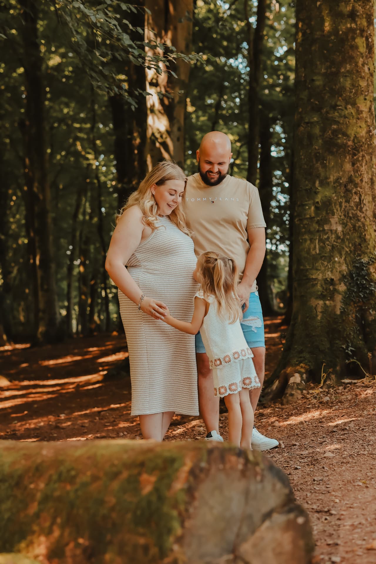 A pregnant mother, her partner and young daughter standing together on a sunlit woodland path