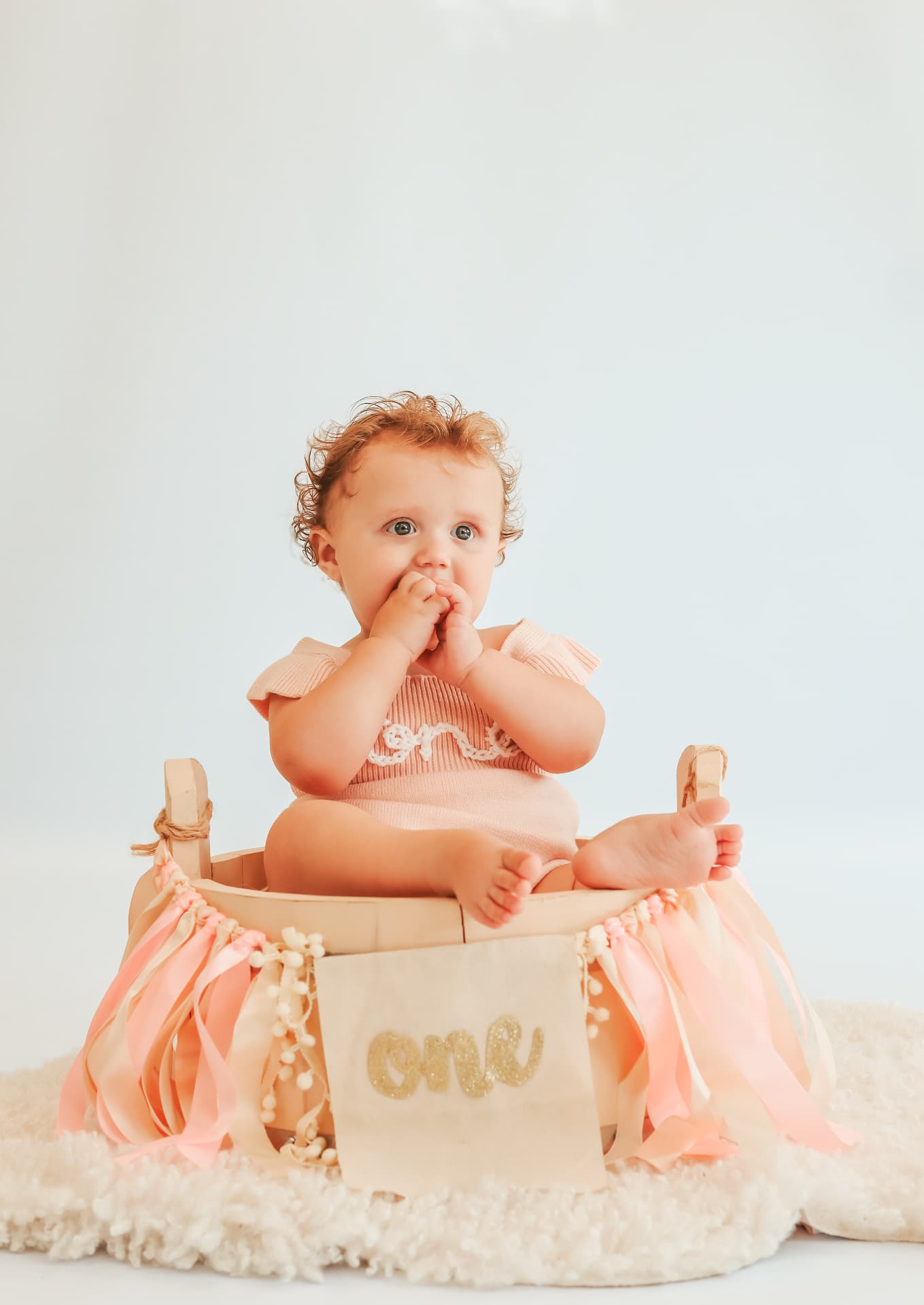 Baby girl sitting in a ribbon-decorated crate with a gold 'one' banner at her first birthday session