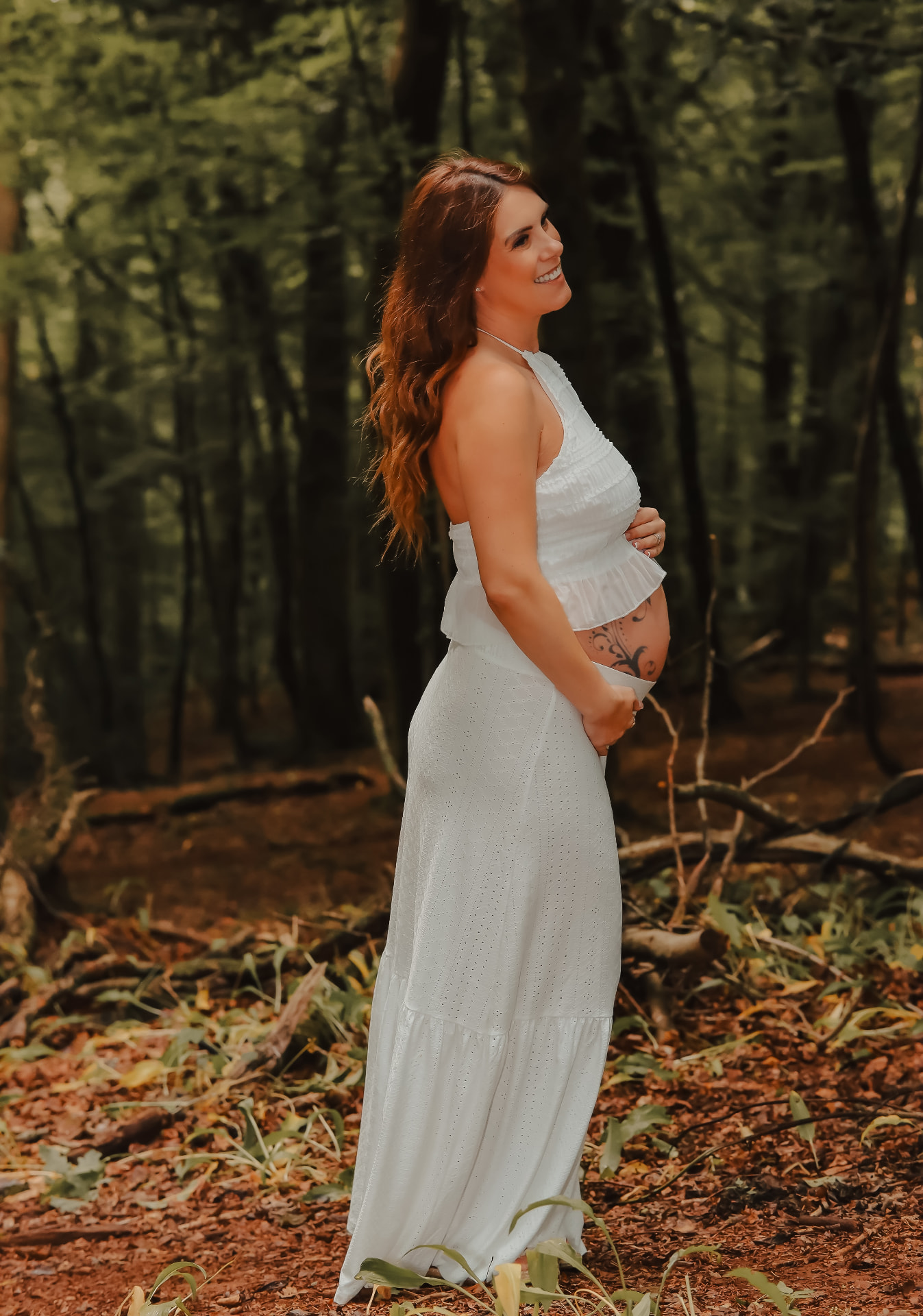 A pregnant woman in a flowing white dress standing in a sunlit woodland