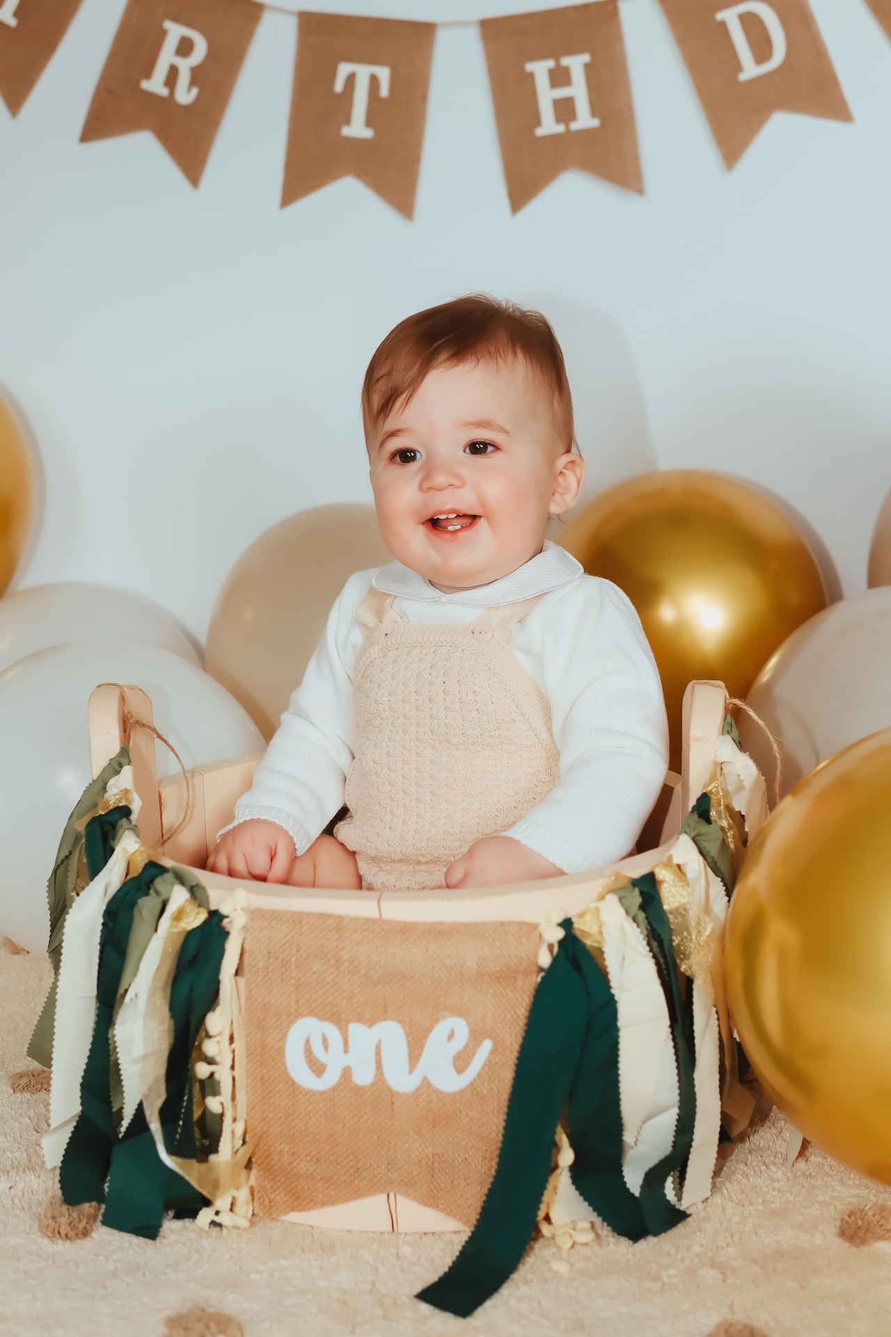 Baby boy in cream knitted dungarees sitting in a decorated crate at his first birthday