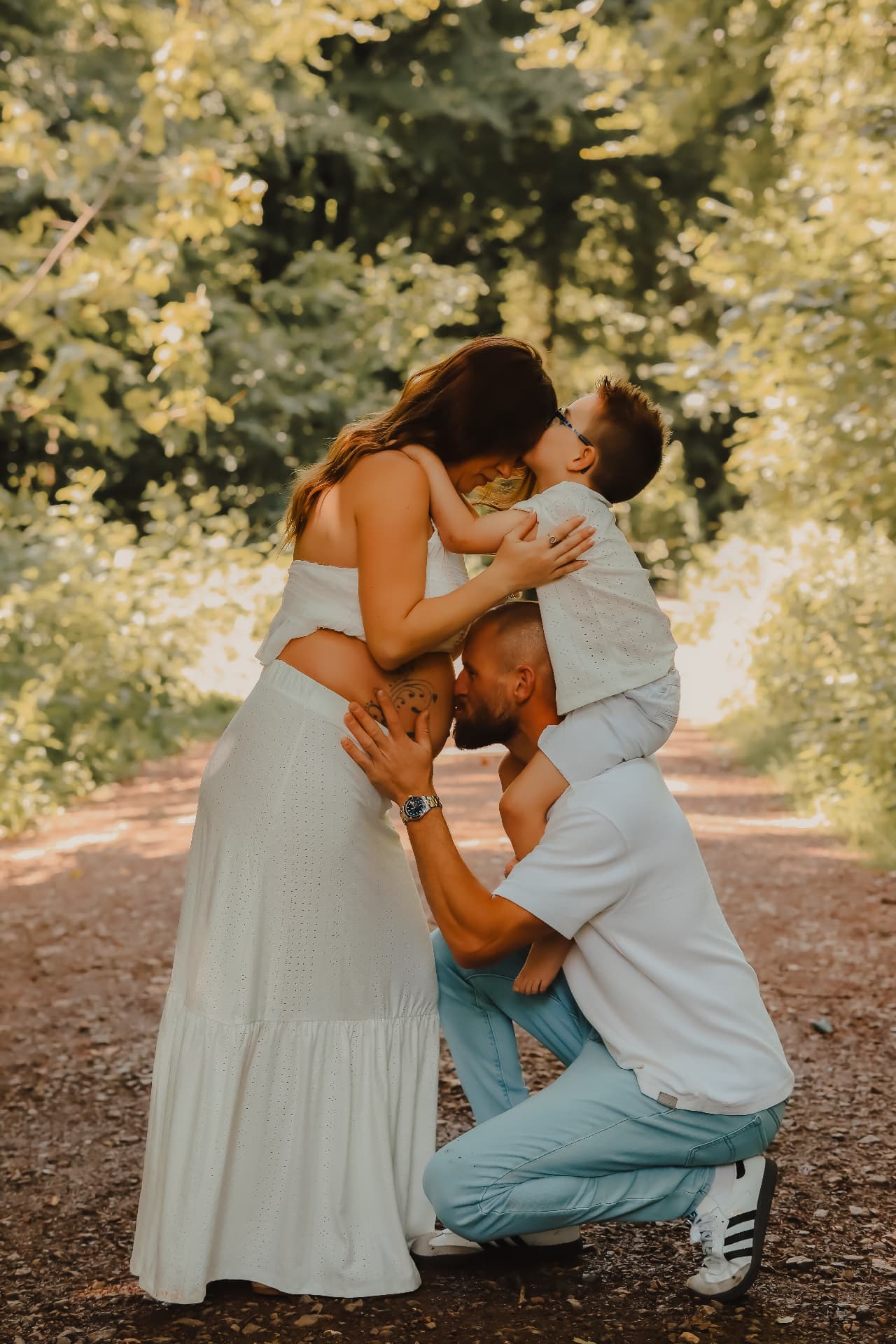 Expectant mother in a white dress held by her partner and young son on a sunlit woodland path