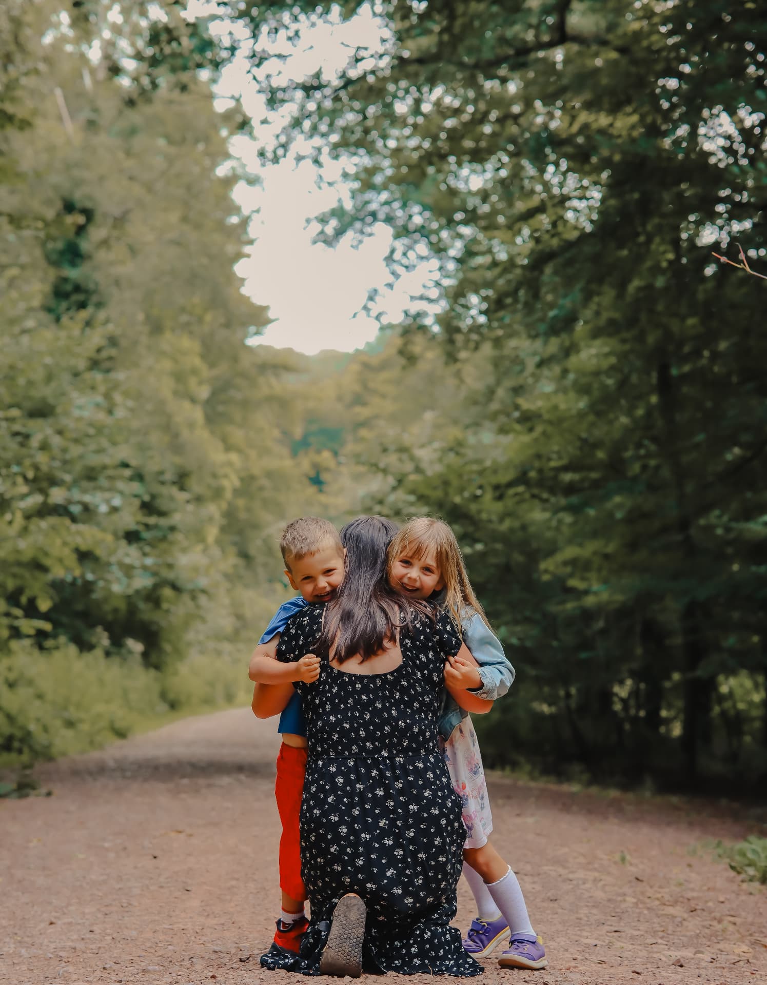 A mother kneeling on a woodland path while two children hug her from behind